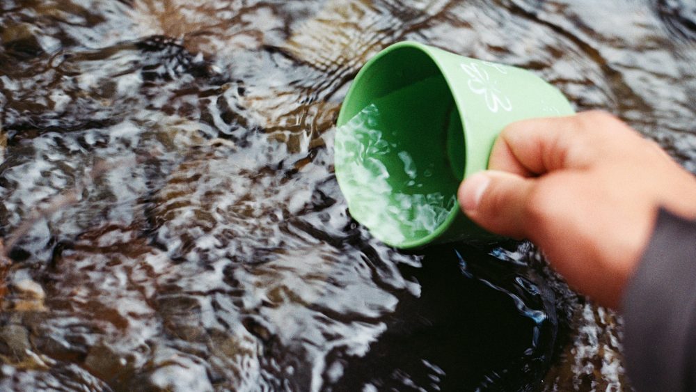 A hand is holding a green cup to scoop up water from a stream