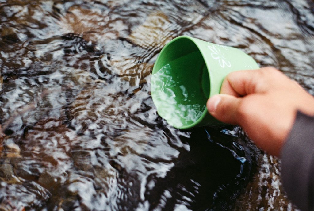 A hand is holding a green cup to scoop up water from a stream