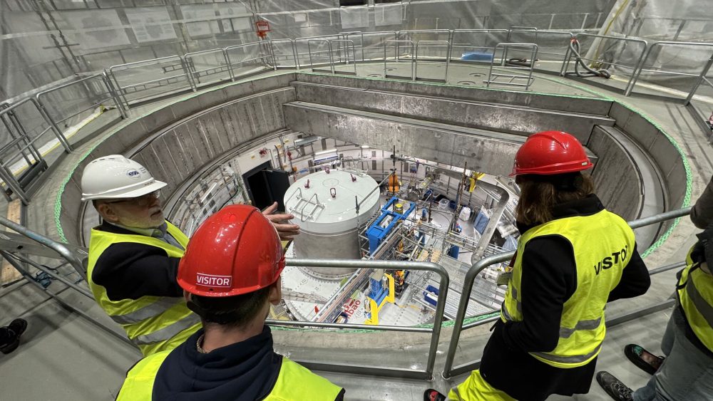 Three people in yellow west and red safety helmets are looking down on advanced equipment at a large scale research infrastructure